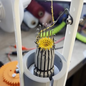 Può includere: Primo piano di una macchina da maglia con un pezzo di uncinetto giallo sugli aghi. La macchina è in plastica bianca e ha una base grigia.