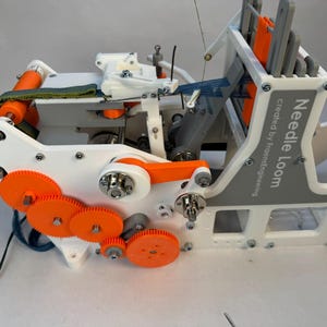 Puede incluir: Un telar de agujas impreso en 3D blanco y naranja con un panel gris que dice "Needle Loom created by FrameKnitting".