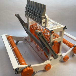 Könnte beinhalten: Ein weißer und orangefarbener 3D-gedruckter Webstuhl mit einer Reihe von grauen Hebeln. Der Webstuhl hat auf jeder Seite eine Spule und eine Reihe von orangefarbenen Stäben.