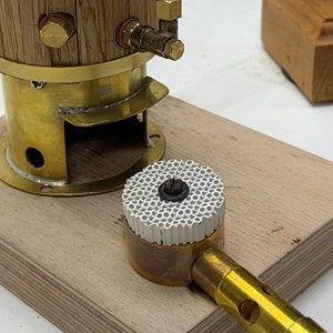May include: A brass steam engine model with a wooden boiler and a ceramic burner. The engine is mounted on a wooden base and is connected to a brass burner with a hose.