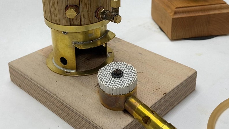 May include: A brass steam engine model with a wooden boiler and a ceramic burner. The engine is mounted on a wooden base and is connected to a brass burner with a hose.