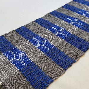 Puede incluir: Un camino de mesa tejido con rayas alternas de gris y azul. Las rayas azules presentan un patr&oacute;n de diamantes blancos.