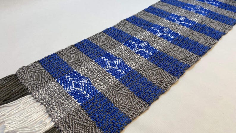 Puede incluir: Un camino de mesa tejido con rayas alternas de gris y azul. Las rayas azules presentan un patr&oacute;n de diamantes blancos.