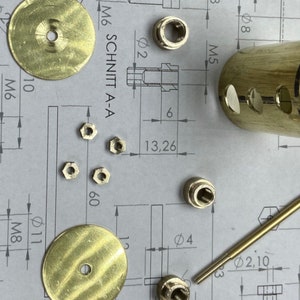 May include: A close-up of brass hardware parts, including two round discs, a cylinder with holes, a long rod, and several small nuts. The parts are arranged on a white sheet of paper with technical drawings and measurements.