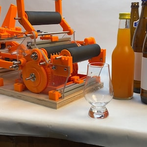 Puede incluir: Una máquina aplicadora de etiquetas naranja impresa en 3D con dos rodillos negros. La máquina está sobre una base de madera. Hay varias botellas de vidrio con etiquetas sobre una superficie blanca delante de la máquina.
