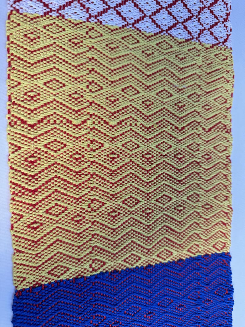 Puede incluir: Una tela tejida con un patr&oacute;n geom&eacute;trico en rojo, amarillo y azul. La tela est&aacute; hecha de un hilo grueso y texturizado.