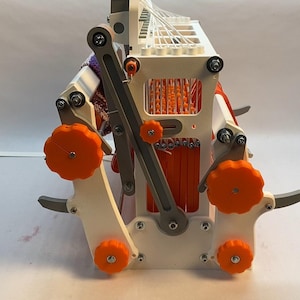 Puede incluir: Un telar blanco impreso en 3D con pomos naranjas y una palanca gris. El telar sostiene una pieza de tejido tejido con rayas naranjas y moradas.