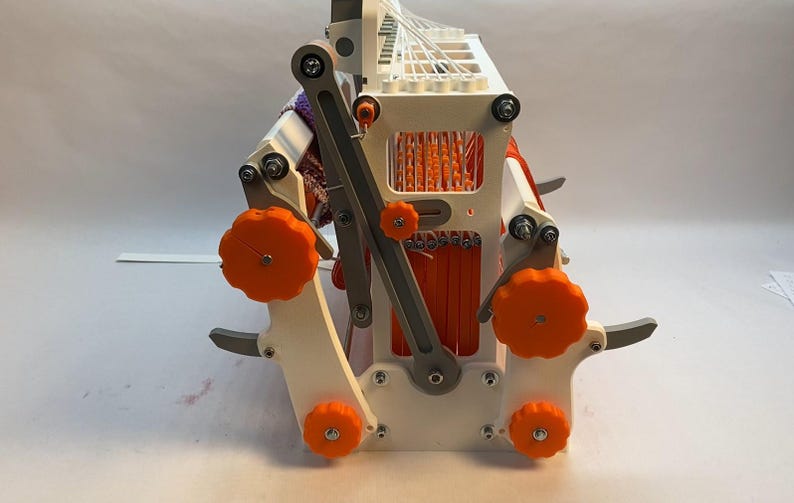 Puede incluir: Un telar blanco impreso en 3D con pomos naranjas y una palanca gris. El telar sostiene una pieza de tejido tejido con rayas naranjas y moradas.