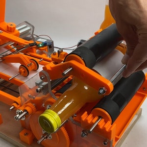 Puede incluir: Una máquina de aplicación de etiquetas de plástico naranja y negro impresa en 3D con una cubierta de plástico transparente. Una mano sostiene un rollo de etiquetas blancas. Una botella de zumo de naranja está siendo etiquetada.