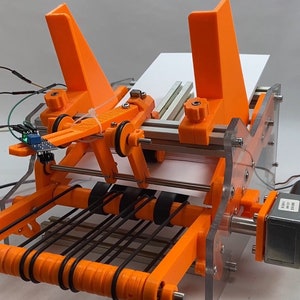 Op de afbeelding: Een oranje 3D-geprinte machine met een transparant kunststof frame en een wit vel papier dat er doorheen beweegt. De machine heeft een motor en een sensor.