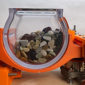 Pode incluir: Um recipiente de plástico transparente cheio de pedras lisas coloridas está montado em uma moldura laranja impressa em 3D. O recipiente gira em uma base com um motor e uma mola.