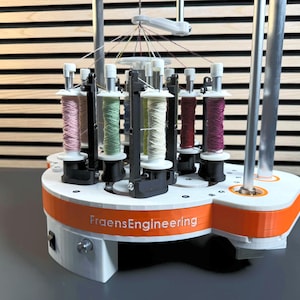 May include: A white and orange FraensEngineering machine with multiple spools of colorful thread. The machine has a central structure with several arms holding the spools, and a base with a power switch. The spools contain pink, green, red, and purple thread.