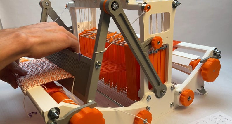 Puede incluir: Un telar impreso en 3D blanco y naranja con hilo naranja enhebrado. Se muestra una mano operando el telar.