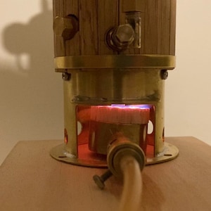 May include: A close-up of a small, brass-colored steam engine with a wooden boiler. The engine is lit and the burner is glowing red.