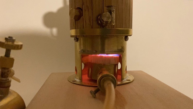 May include: A close-up of a small, brass-colored steam engine with a wooden boiler. The engine is lit and the burner is glowing red.