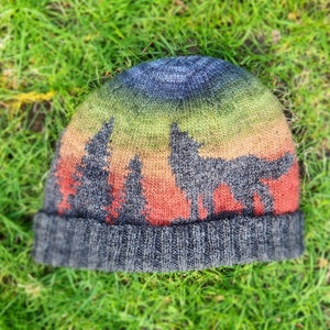 Puede incluir: Un gorro de punto con un diseño degradado de colores que presenta una silueta de un lobo y árboles. El gorro es gris con un borde gris oscuro.