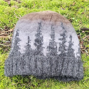 Könnte beinhalten: Eine graue und braune Strickmütze mit einem Waldmotiv. Die Mütze hat einen Umschlag und ist aus einem weichen, warmen Material gefertigt.