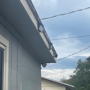 May include: Exterior view of a house with light blue cladding and a grey roof. Several black security lights are mounted along the roofline. A white security camera is mounted on the side of the house. The sky is overcast.