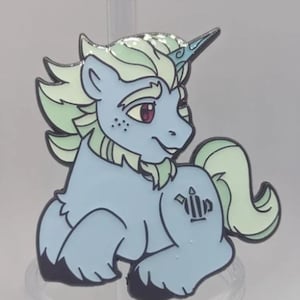 Könnte beinhalten: Hellblaue Einhorn-Emaille-Anstecknadel mit grüner Mähne, Schweif und Akzenten. Das Einhorn hat ein lila Auge und ein Horn. Die Anstecknadel hat eine schwarze Umrandung und ein Tattoo am Arm. Ein Sammlerstück.