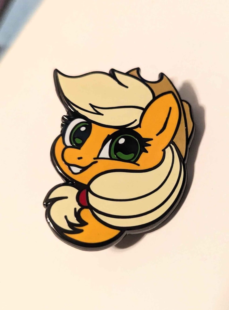 Peut inclure: Une &eacute;pingle en &eacute;mail jaune et blanche repr&eacute;sentant un poney de dessin anim&eacute; avec un chapeau marron et des yeux verts. Le poney sourit et a un c&oelig;ur rouge sur la poitrine.