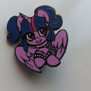 Könnte beinhalten: Eine Emaille-Anstecknadel mit einem Cartoon-Pony mit rosa Mähne und Schweif, das eine schwarze Halskette und Armbänder trägt. Das Pony hat ein rosa Horn und blaue Augen.