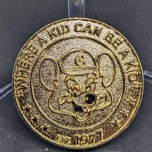 Puede incluir: Una moneda dorada con la frase "WHERE A KID CAN BE A KID" alrededor del borde. La moneda presenta un ratón de dibujos animados con un sombrero y el texto "EST. 1977".