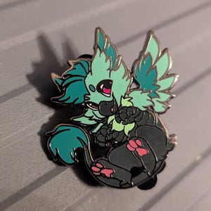 Op de afbeelding: Een teal en zwarte emaille pin met een klein, gevleugeld wezen met een pluche staart en roze pootjes. Het wezen is afgebeeld in een speelse pose, met zijn vleugels uitgespreid en zijn hoofd schuin.
