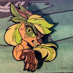 May include: A green and yellow enamel pin featuring a cartoon animal with a fierce expression. The animal has a leafy crown and a long mane of yellow hair.