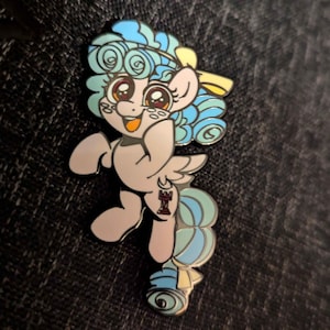 Op de afbeelding: Een pastelblauwe en gele emaille pin met een cartoon pony met een gekrulde manen en staart. De pony draagt een gele strik en heeft een roze neus en een rood-zwart schaakstuk op zijn been.