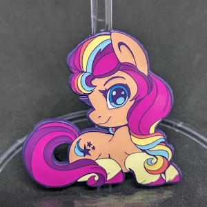 Könnte beinhalten: Eine lila, rosa und gelbe Ponyfigur mit einer Regenbogenmähne und einem Regenbogen-Schwanz. Das Pony hat ein sternförmiges Tattoo an der Seite und ein blaues Auge.