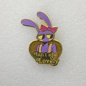 Könnte beinhalten: Eine violette Emaille-Anstecknadel mit einem Cartoon-Hasen mit langen Ohren, einer rosa Schleife und gelben Augen. Der Hase ist vor einem goldglitzernden Herz platziert, auf dem der Text Isn't she Lovely in einer goldenen Schreibschrift steht.