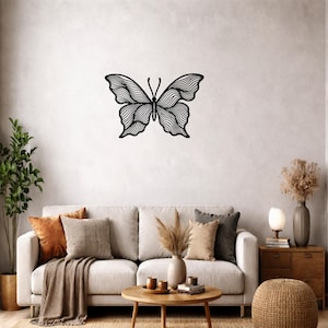 Puede incluir: Una pieza de arte mural de metal negro con forma de mariposa que presenta intrincados patrones de líneas onduladas y fluidas en sus alas. La mariposa está montada en una pared lisa de color claro sobre un sofá, con un diseño simétrico y dos antenas.