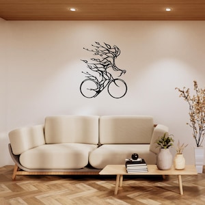 Puede incluir: Arte mural de metal negro que representa una bicicleta con un ciclista, estilizado con líneas fluidas y detalles en forma de hojas. La obra de arte está montada en una pared blanca sobre un sofá beige.