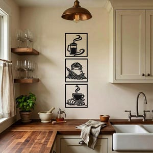 Puede incluir: Una pieza de arte mural vertical que consta de tres marcos cuadrados negros apilados. El marco superior muestra una cafetera de émbolo, el central representa un saco de granos de café y el inferior presenta una taza de café con un croissant.