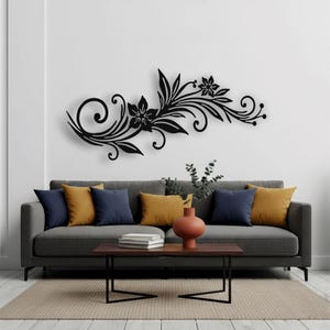 Puede incluir: Una pieza de arte mural floral de metal negro con un diseño fluido de hojas, flores y espirales, montada sobre un sofá gris con cojines amarillos y azul marino. Una mesa de centro marrón y un jarrón de terracota están frente al sofá.