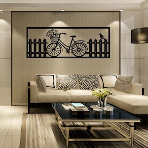 Puede incluir: Arte mural de metal negro que representa una bicicleta con una cesta de flores, una valla y un pájaro. La obra de arte rectangular está montada en una pared beige sobre un sofá crema. La habitación tiene un diseño moderno.
