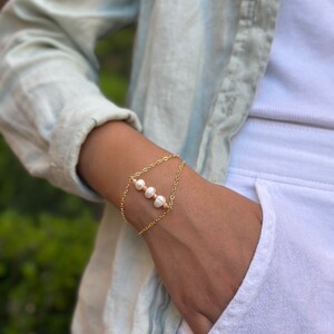 May include: A gold chain bracelet with a delicate design featuring four white pearls. The bracelet is layered with two chains, creating a stylish and elegant look.
