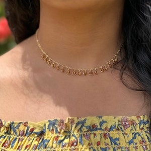 May include: A gold chain necklace with small, round, brown beads dangling from it. The necklace is worn by a person with dark hair and a yellow floral top.