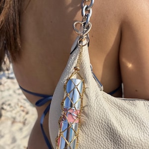 May include: A cylindrical lipstick tube encased in a decorative gold-toned metal mesh net holder. The holder is attached to a silver-toned chain strap of a white textured handbag by a small heart-shaped clasp, featuring a light pink lipstick shade.