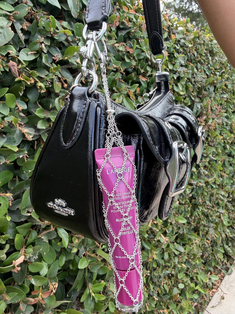 May include: A black patent leather Coach shoulder bag with a silver-toned metal chain harness holding a magenta Laneige Lip Glowy Balm tube. The bag features a front flap with a silver buckle and a silver logo on the side. The lip balm tube is labeled Blueberry.