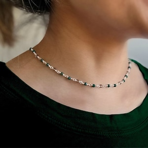 May include: A delicate necklace featuring small, dark green beads interspersed with clear beads, strung on a silver chain. The necklace is worn around the neck, creating a subtle and elegant accessory. The dark green beads provide a pop of color against the silver chain.
