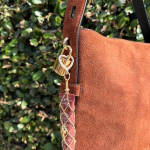 May include: A brown suede shoulder bag featuring a gold-tone heart-shaped padlock charm attached to the strap. A small tube of Laneige lip product is encased in a decorative gold-tone metal chain mesh holder, suspended from the padlock by a chain.