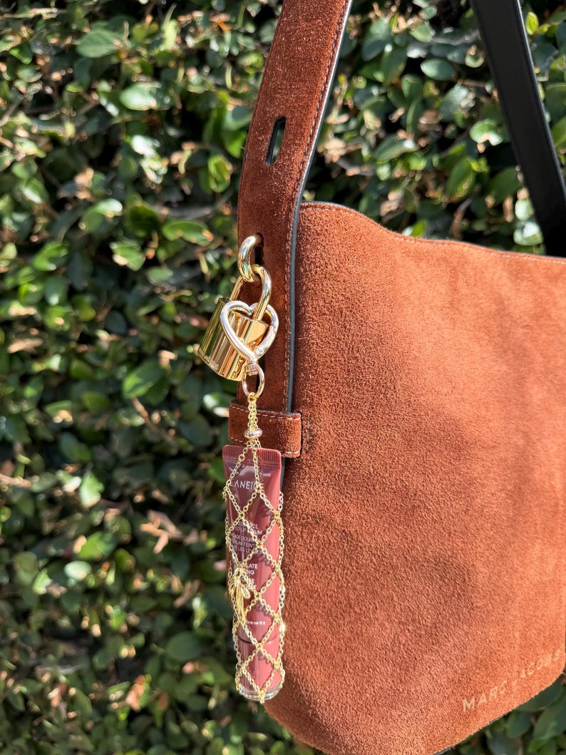 May include: A brown suede shoulder bag featuring a gold-tone heart-shaped padlock charm attached to the strap. A small tube of Laneige lip product is encased in a decorative gold-tone metal chain mesh holder, suspended from the padlock by a chain.