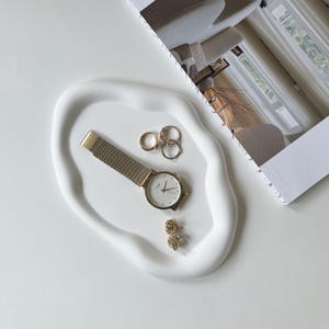 Pode incluir: Uma bandeja branca de formato orgânico contém um relógio de pulso dourado, vários anéis de ouro e dois pares de brincos de ouro. Uma revista com uma foto de um quarto está no fundo. Os itens estão dispostos em uma superfície branca.