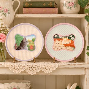 May include: Two round ceramic decorative plates displayed on small gold stands. The left plate features a grey cat in a garden, while the right plate shows three light-colored kittens in a basket. Both plates have patterned borders.