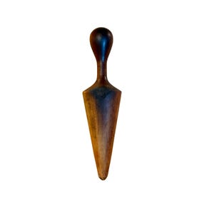 May include: A dark brown wooden tailor's clapper with a smooth, rounded handle at the top and a wide, tapered body that narrows to a point at the bottom. The wood grain is visible throughout the surface, showing variations in color and texture.