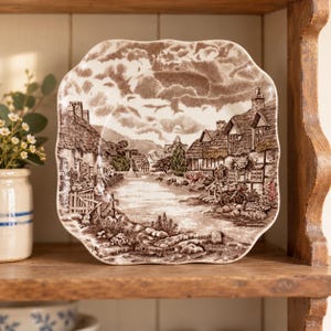 May include: A square ceramic plate with scalloped edges featuring a brown and white transferware print of a rural village scene with thatched-roof cottages, a central path, and detailed foliage under a cloudy sky, presented in a classic style.