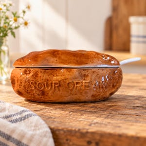 May include: A brown, potato-shaped ceramic crock with a matching lid and a white spoon. The word SOUR CREAM is embossed in capital letters on the front of the container. The surface has a mottled, textured finish resembling a baked potato.