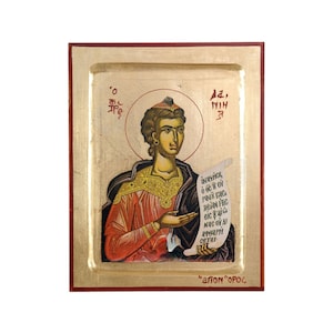 May include: A hand-painted icon of a saint holding a scroll with Greek text. The icon is framed in a gold-coloured frame with a red border. The text on the scroll reads "ΜΗΝΥΣΗΣ Ο ΘΕΟΣ ΣΟΥ ΠΑΥΣ ΕΣΘΕ ΔΕΙΛΙΑΣ ΗΣ ΕΙΣ Σ' ΑΥΤΟΣ ΝΑΘ ΟΙ ΑΒΒΑΡΙΗ ΣΕΤΙ". The text at the bottom of the icon reads "ΑΓΙΟΝ ΟΡΟΣ".