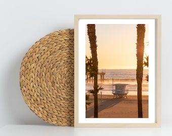 May include: A framed photograph of a beach scene with a lifeguard stand, a pier, and palm trees in the foreground. The sky is a vibrant orange and yellow, creating a warm and inviting atmosphere.
